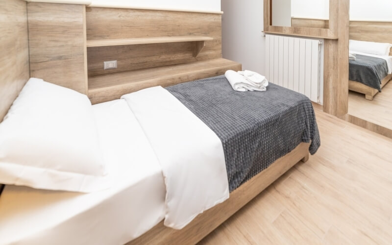 Hotel room with two beds, one angled in foreground, white sheets, dark blanket, and minimalist decor reflected in a mirror.