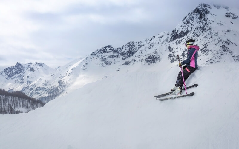 Skier in pink and gray jacket mid-air over snow-covered mountain, with majestic peaks and cloudy sky in the background.