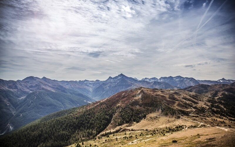 Paesaggio montano con cielo nuvoloso, sentieri di terra e colline coperte di alberi e erba, che suggeriscono un ambiente sereno.