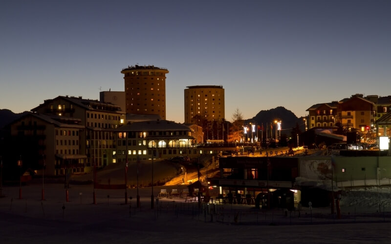 Cityscape at dusk with illuminated buildings, a deep blue sky, and mountains in the background, creating a serene atmosphere.