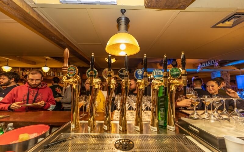 Lively bar scene with a row of beer taps, patrons enjoying drinks, warm lighting, and wooden accents creating a cozy atmosphere.