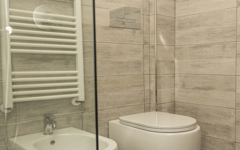 Compact bathroom with a glass door, sink, toilet, heated towel rail, and light gray tiled walls.