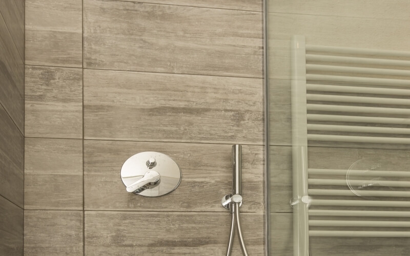 Shower wall with light brown tiles, silver temperature knob, handrail, and vent, showcasing simplicity and elegance.