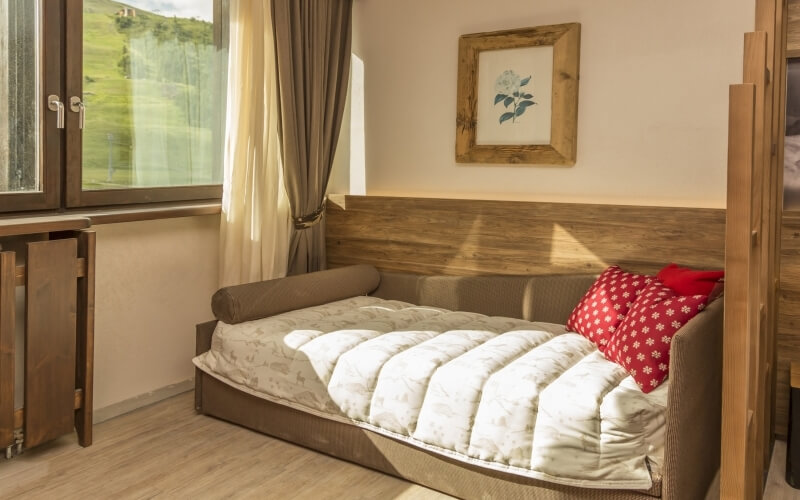 Cozy room with a daybed, brown couch, red pillow, natural light, white walls, and a view of a green hillside.