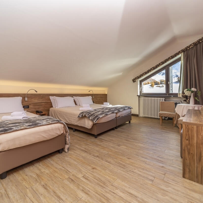 Cozy room with two beds, a window with curtains, and a table with flowers, featuring white walls and light wood flooring.