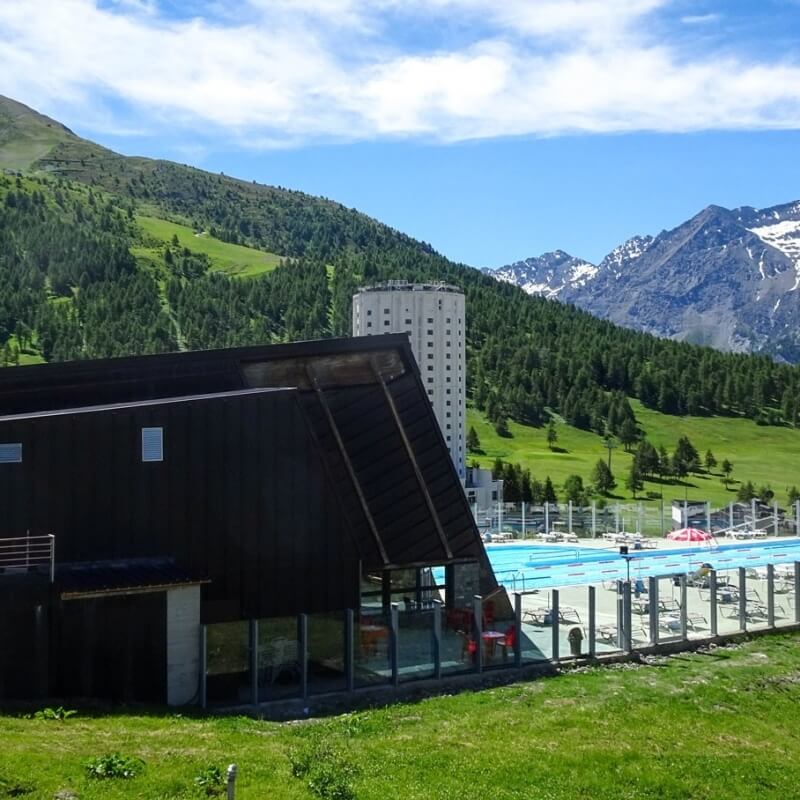 Serene mountain scene with a pool, lounge chairs, and buildings, under a blue sky with clouds and lush green mountains.