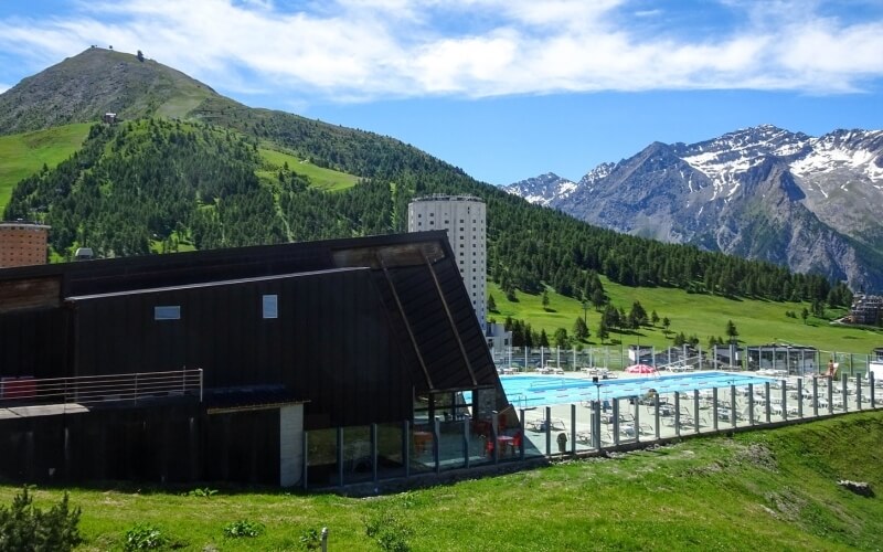 Outdoor swimming pool in the mountains, surrounded by a safety fence, with an A-frame building and lush greenery.