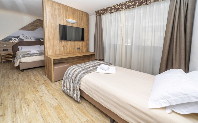 Hotel room with two beds, a mounted TV, window with curtains, and light wood flooring, creating a serene atmosphere.