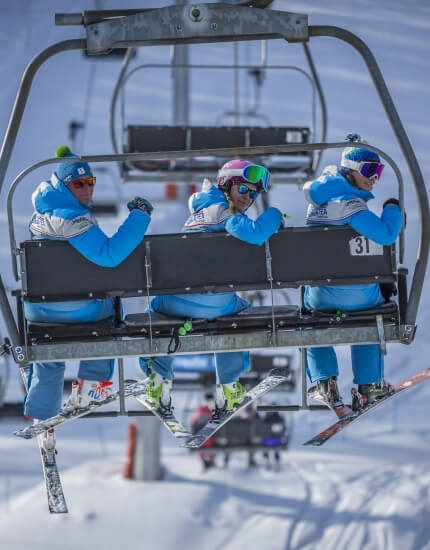 Three skiers in blue and white uniforms ride a chairlift at a snowy ski resort, with more chairlifts in the background.