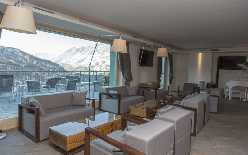 Modern open-plan living space with gray furniture, white piano, mountain view, and patio area through large window.