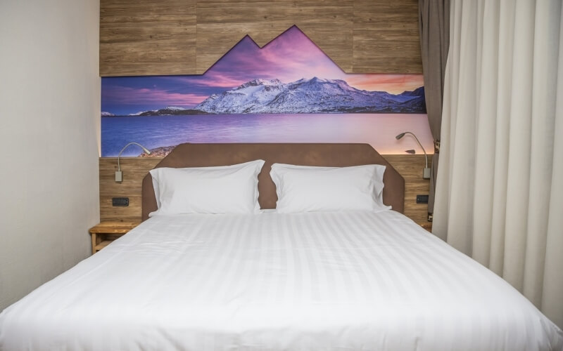 Serene bedroom with a large bed, white linens, vibrant mountain mural, and soft off-white walls, creating a tranquil atmosphere.