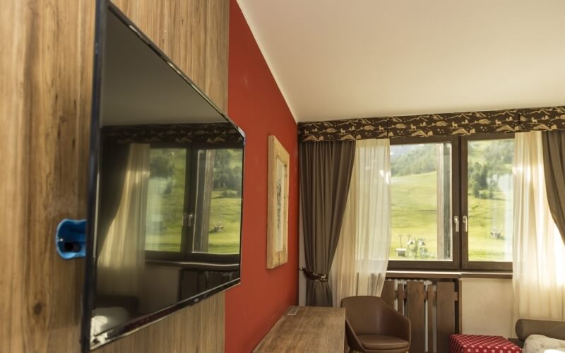 Cozy room with a mounted TV on a red wall, a large window with curtains, a brown chair, and a bed with a checkered blanket.