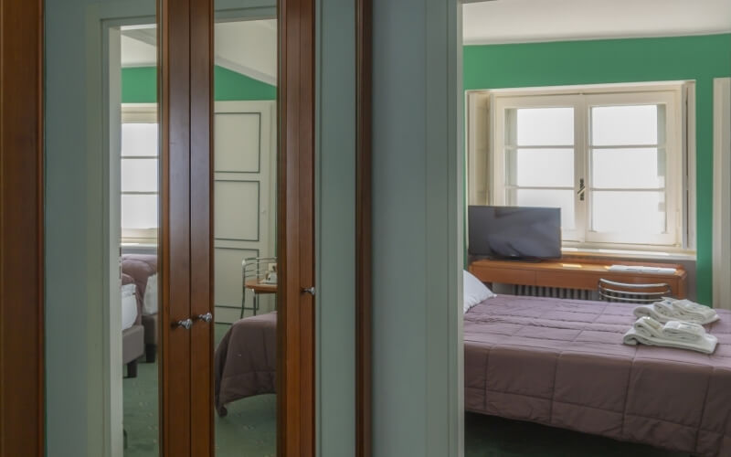 A serene green bedroom with a bed, desk, and television, featuring a pink comforter and white windows.