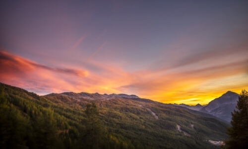 Serene mountainous landscape at sunset with a purple sky, lush foreground, distant mountains, and a valley with buildings.