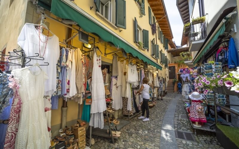 Charming cobblestone street with yellow shops, green awnings, and shoppers browsing vibrant merchandise.