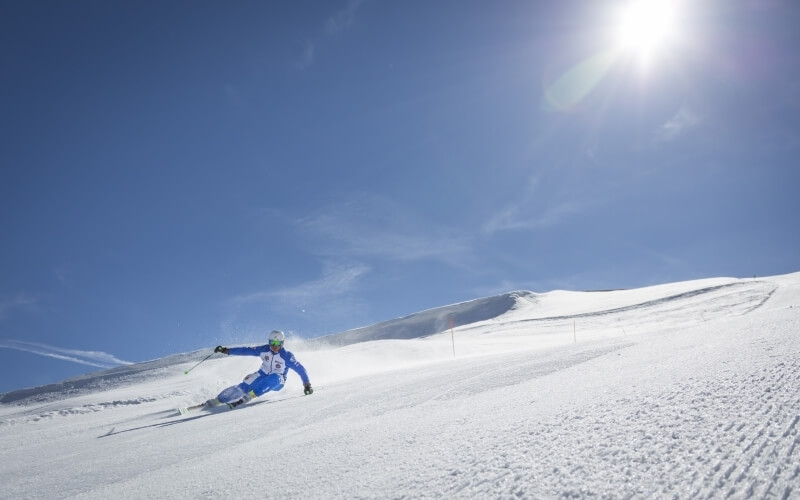 Skier in blue and white gear glides down a sunny slope, surrounded by snow and a bright blue sky with clouds.