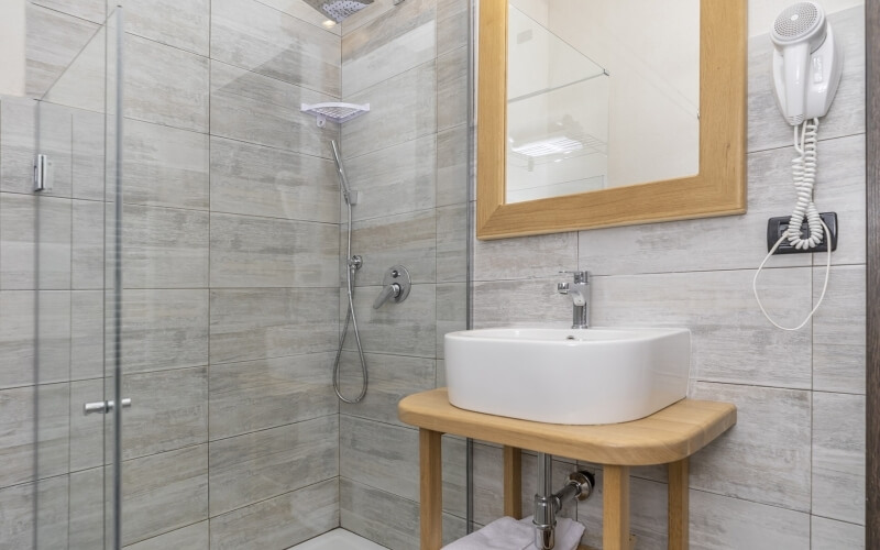 A modern bathroom featuring a wooden mirror, white sink, hair dryer, glass shower door, and gray tiled walls.