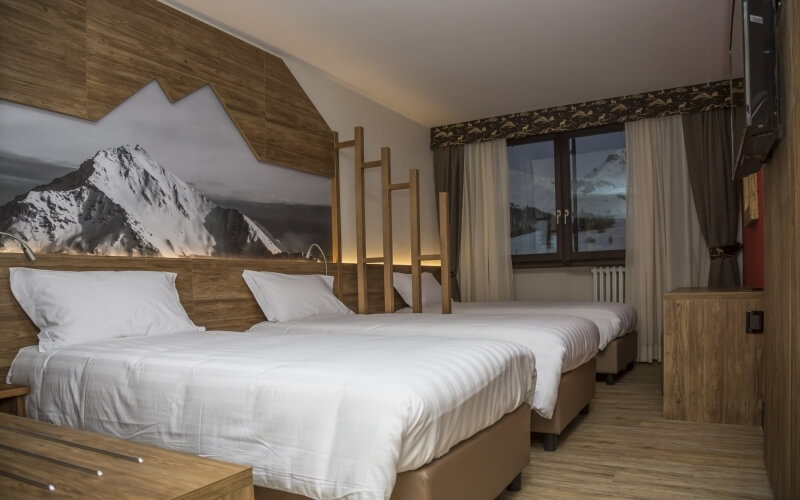 Cozy hotel room with two beds, mountain mural, TV, and window view of snow-capped mountains, decorated in rustic style.