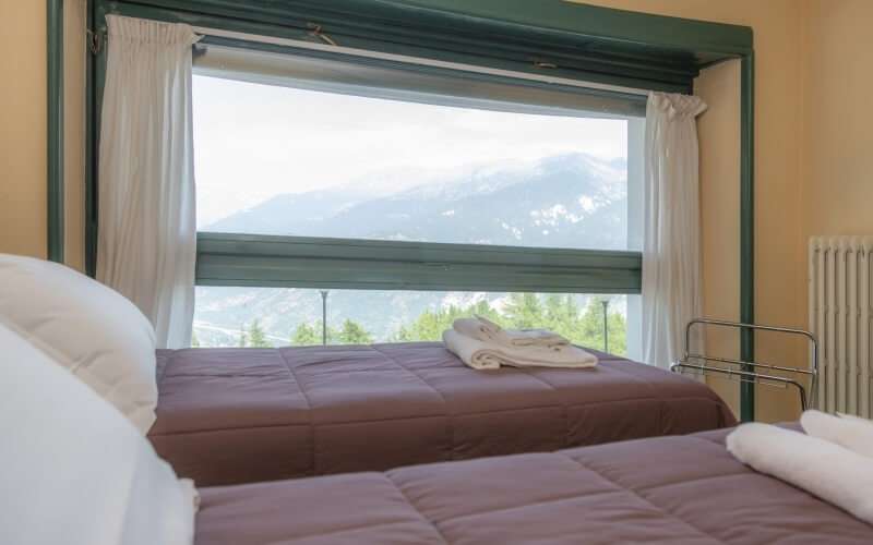 Serene bedroom with two beds, yellow walls, and a large window showcasing majestic mountains and a bright sky.