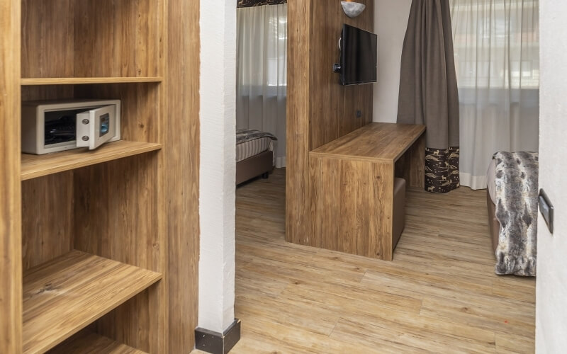 Cozy room with wooden floor, desk and TV, bed with patterned blanket, shelving unit, and white curtains.