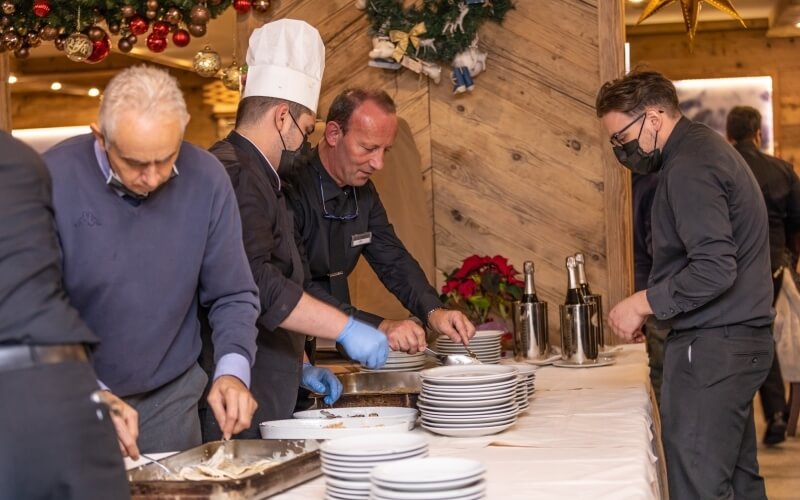 A group of men in various attire serve food at a holiday event, with a chef overseeing from a table covered in white cloths.