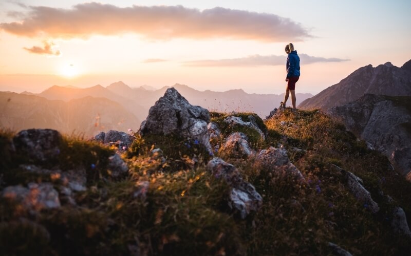 Una donna in giacca blu e pantaloni rossi osserva un maestoso paesaggio montano al tramonto da un affioramento roccioso.