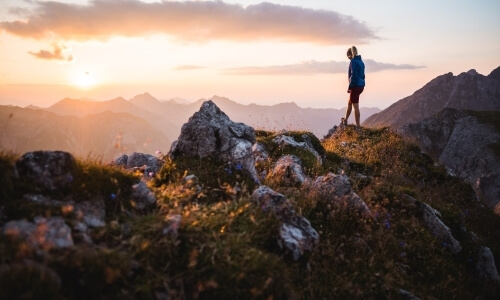 Two individuals stand on a rocky outcropping, surrounded by greenery, against a vibrant sunset over majestic mountains.