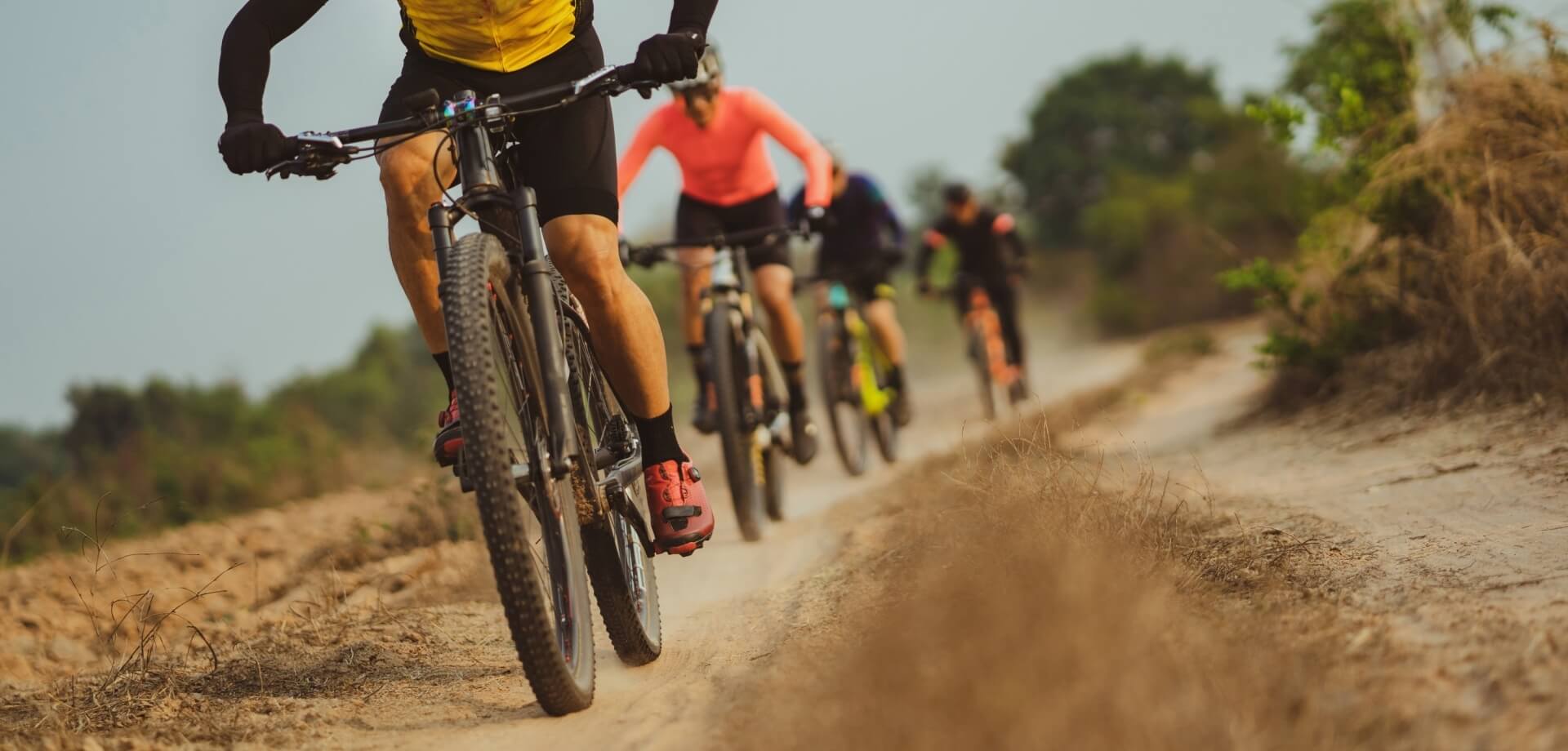 A group of cyclists rides single file on a dirt path through a rural area, led by a cyclist in a yellow shirt.