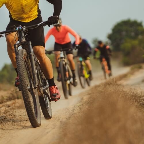 A group of cyclists rides on a dirt path under a blue sky, with a prominent cyclist in a yellow shirt in the foreground.