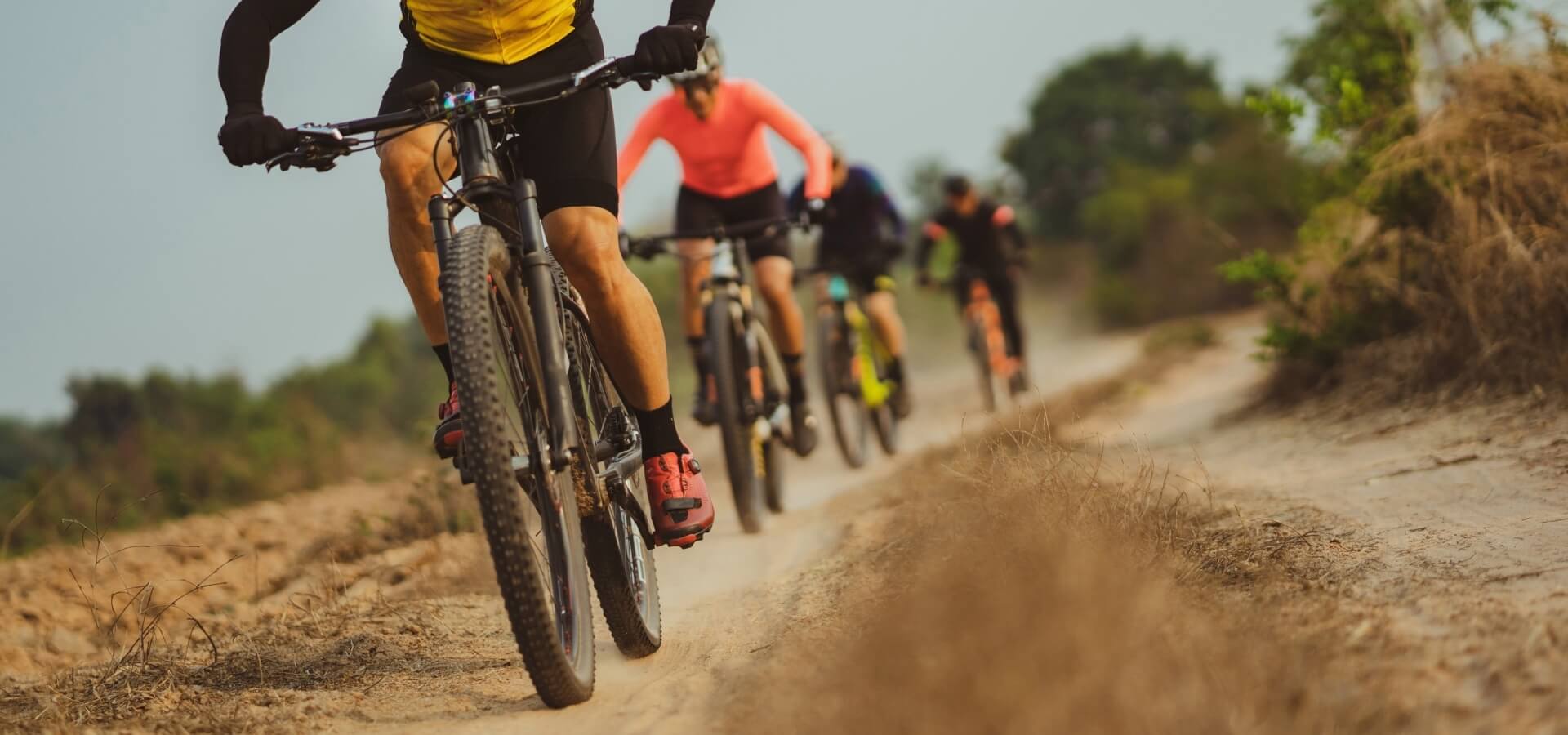 A group of cyclists in athletic gear rides mountain bikes on a dirt path surrounded by trees under a gray sky.