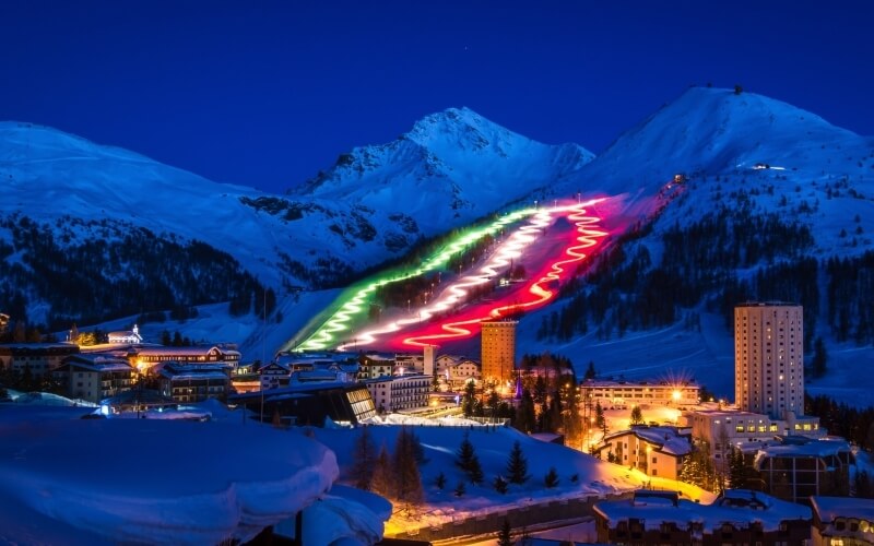 Snow-covered mountains under a dark blue sky, featuring a ski slope with Italian flag lights and illuminated buildings in the foreground.