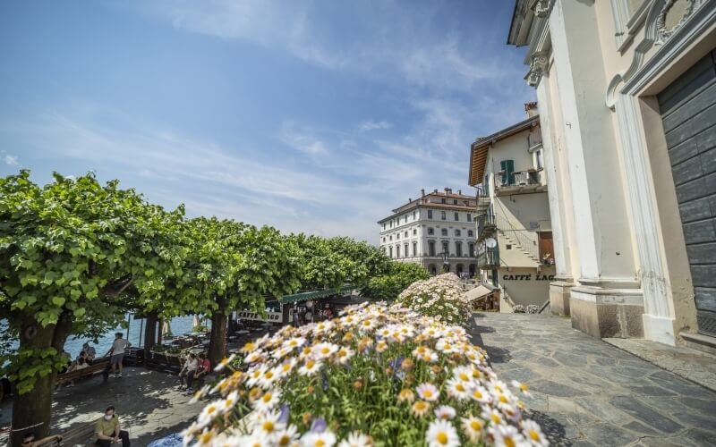 Picturesque European street with cobblestones, balconies, trees, white flowers, outdoor cafes, and a "Caffè Lac" sign.