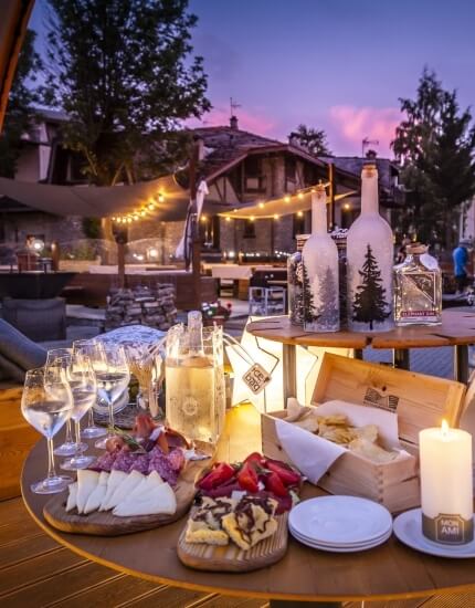 Outdoor round table with cheeses, pastries, wine, and a candle, set against a stone building and trees under a blue-pink sky.