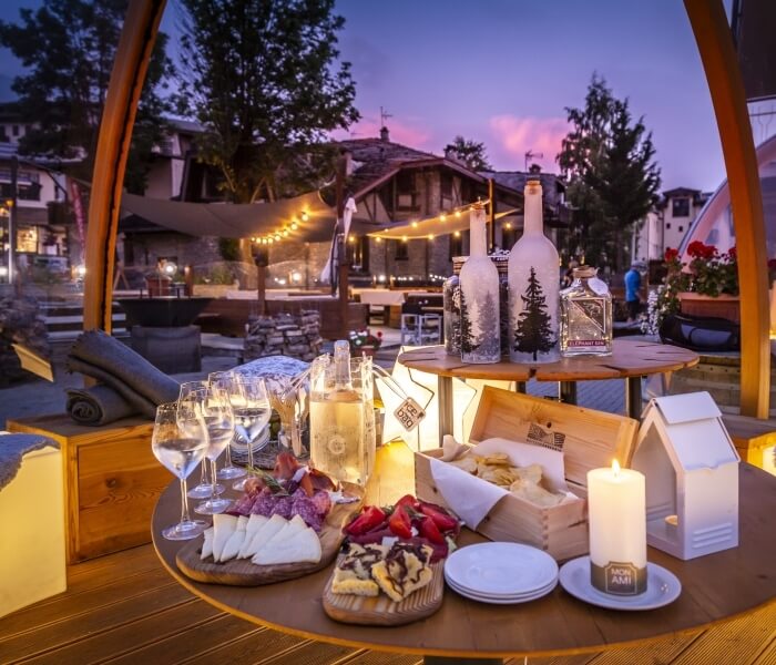 Outdoor table with food and drinks, surrounded by greenery, a cozy building, and a soft pink sky at sunset.