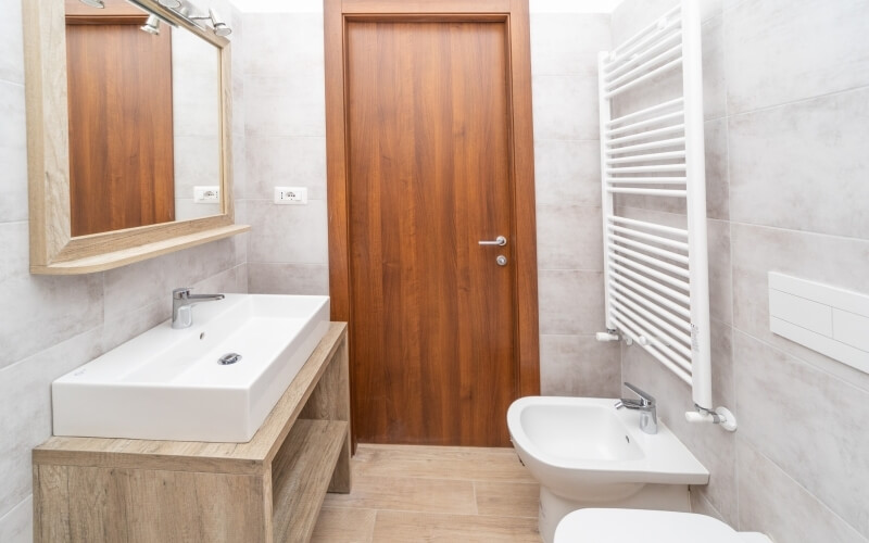 Modern bathroom featuring a white sink, toilet, large mirror, wooden vanity, and light-colored tiled walls and floor.