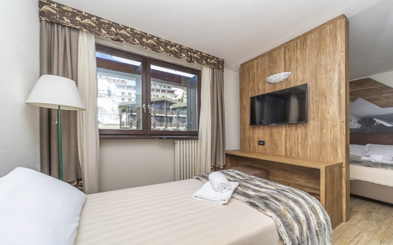 Cozy hotel room with two beds, a TV, window with curtains, and a lamp beside a bed with a striped blanket and towel.