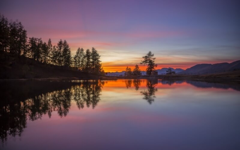 Paesaggio sereno con acqua calma, alberi, montagne e cielo vibrante al tramonto, riflessi armoniosi sulla superficie.