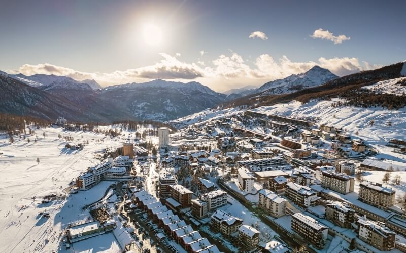 Bird's-eye view of a snow-covered town in mountains at sunrise/sunset, with skiers' paths and a partly cloudy sky.