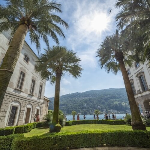 Serene courtyard with palm trees, individuals walking, and a view of a glacial blue lake and mountains under a radiant sky.