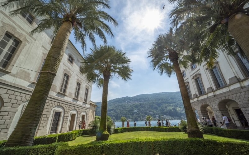 Serene lakeside scene with a white stone building, palm trees, a manicured lawn, and distant rolling hills under a blue sky.