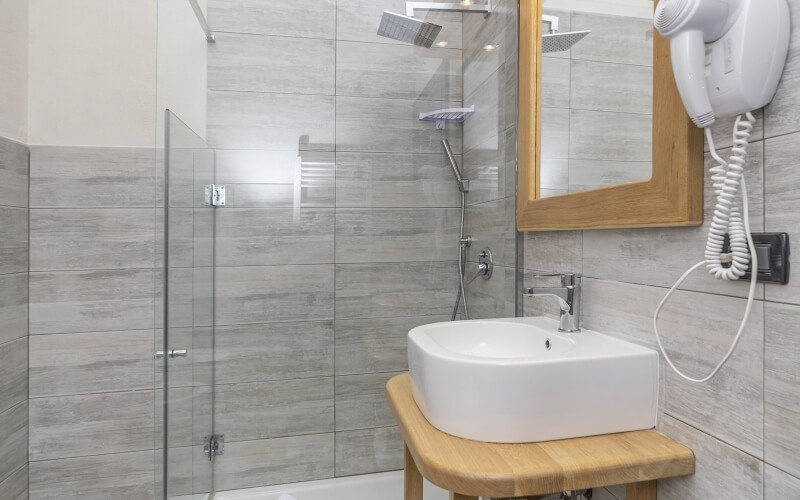 A modern bathroom featuring a glass shower, white sink on a wooden counter, mirror, and wall-mounted hair dryer.