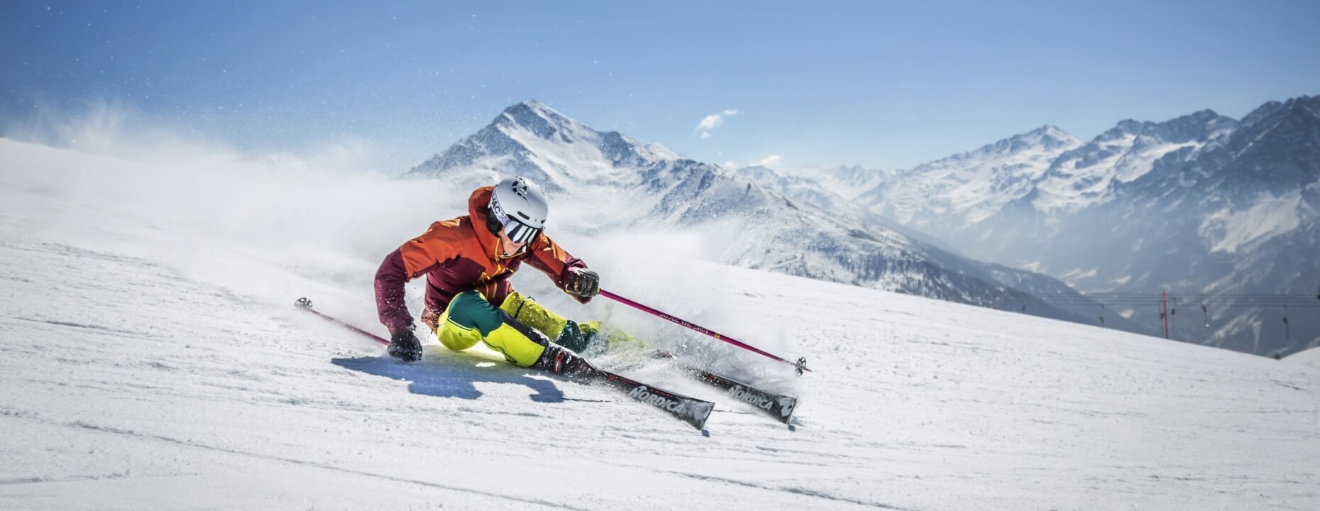 Skier in white helmet and orange jacket descends snowy slope against a clear blue sky and mountain range.