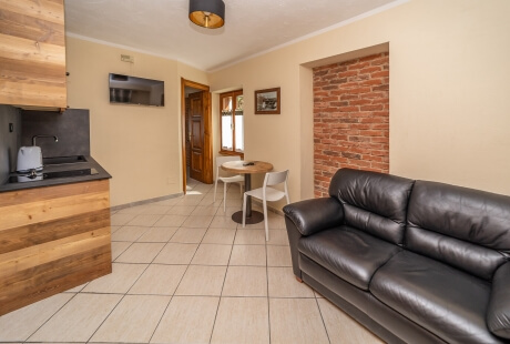 A cozy living room with a kitchen, featuring beige tones, wood accents, a red brick wall, and a leather couch.