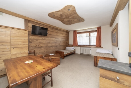 Camera con tavolo e sedie in legno, due letti, TV a parete e finestra con tende rosse, ben illuminata e accogliente.