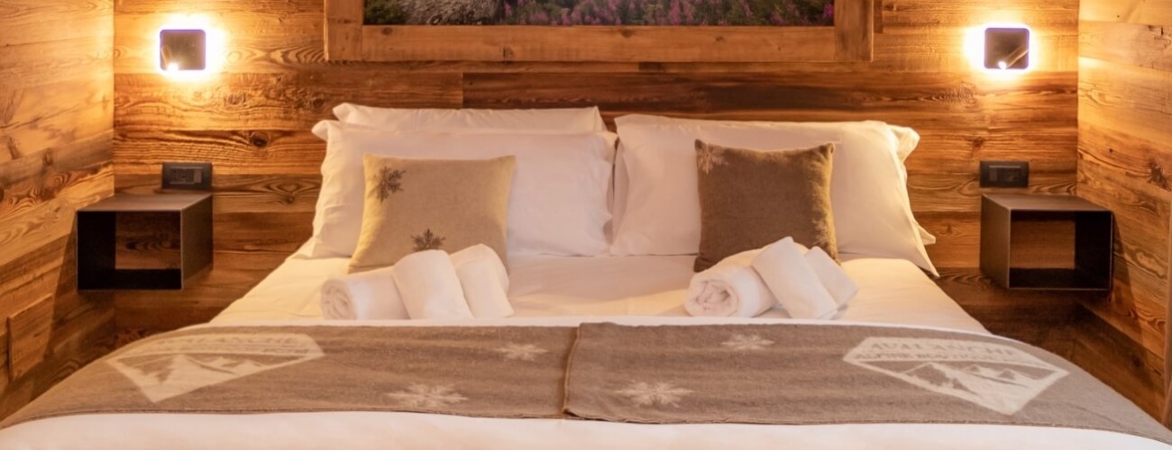 A bed with white linens and pillows, a brown blanket, wooden wall, and nightstands with wall lighting and artwork above.