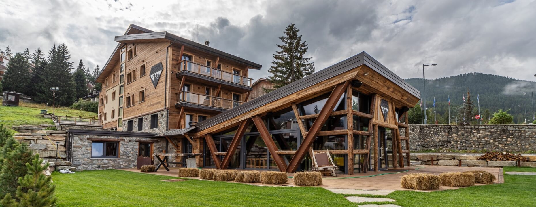 Modern rustic building with wood and stone, surrounded by lush greenery and a dense forest under an overcast sky.