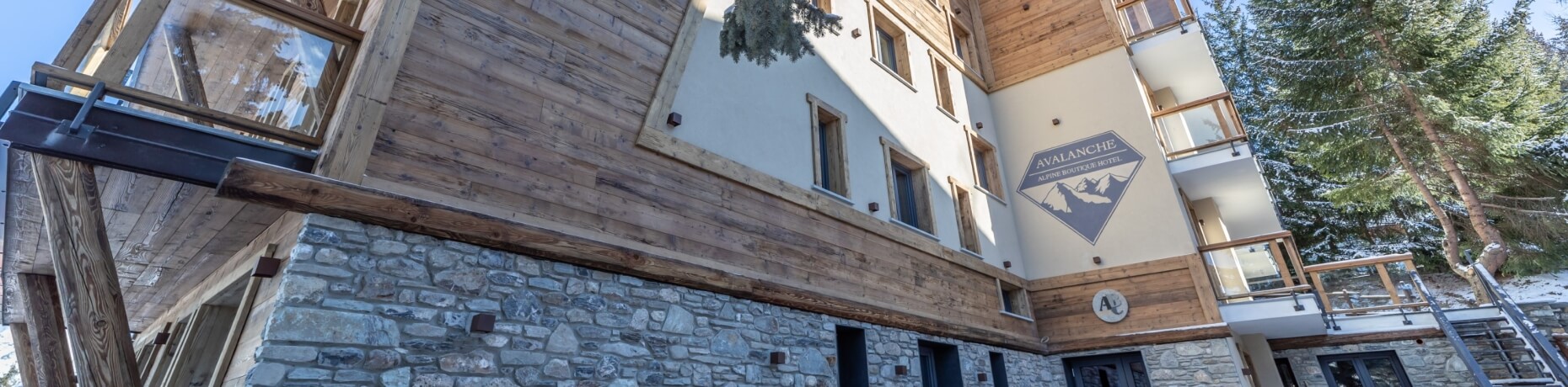 Three-story alpine boutique hotel with stone base, wooden balconies, and mountain sign, surrounded by snow-covered trees.