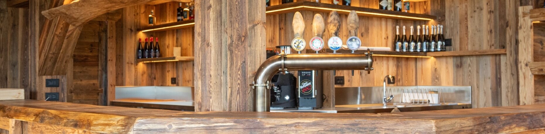 Bar in legno rustico con rubinetto metallico e scaffali illuminati per vini, creando un'atmosfera accogliente e autentica.