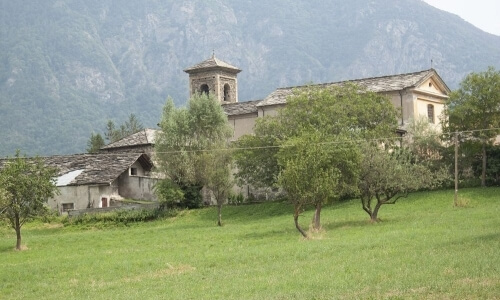 Chiesa pittoresca in una valle tranquilla, circondata da montagne e alberi, con un sentiero di terra che conduce all'ingresso.