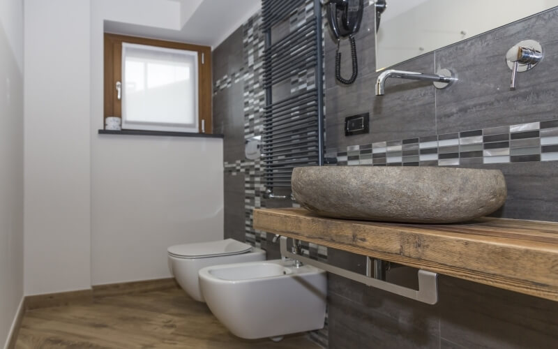 Modern bathroom with stone sink, wooden countertop, two toilets, gray and white tiles, and natural light from a window.